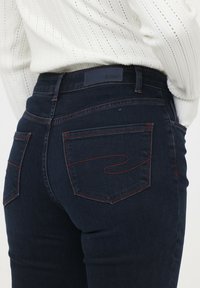 Vaqueros de mezclilla azul oscuro con cintura alta, sutiles bolsillos traseros y costuras rojas en contraste. Textura suave con un diseño ajustado.