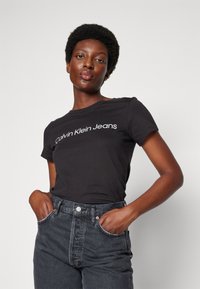 Černé tričko s krátkým rukávem s bílým logem "Calvin Klein Jeans". Spárované s tmavě šedými džínami s vysokým pasem, které mají kapsy.