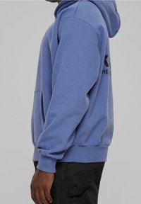 Ljusblå hoodie med en känguruficka, ribbade ärmslut och nederkant. Har en djärv svart logga på baksidan. Mjuk tygkänsla.