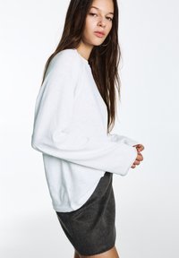 Camicia bianca a maniche lunghe con vestibilità rilassata e polsini larghi, abbinata a una gonna grigio scuro. Tessuto dalla texture liscia, design minimalista.