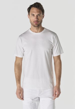 Hombre con pelo corto y barba, vistiendo una camiseta blanca de manga corta lisa y pantalones blancos, de pie frente a un fondo gris claro.