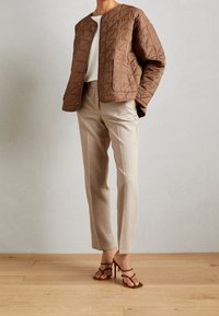 Quilted brown jacket with round neckline, paired with beige tailored pants. The model wears brown strappy heels. Smooth texture, modern design.