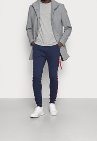 Grau Wollmantel, hellgraues T-Shirt, marineblaue Jogginghose mit roten Seitenstreifen und weiße Sneaker. Slim Fit mit einer Reißverschlusstasche.