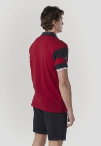 Polo rouge avec col, présentant des rayures noires horizontales sur les manches et un écusson logo. Confectionné en tissu lisse et respirant.