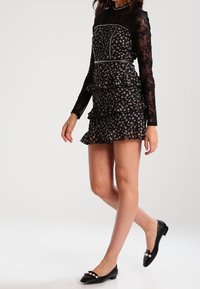 Schwarz floral bedrucktes Kleid mit langen Spitzenärmeln, gerafftem Rock und Perlenverzierungen an der Taille. Schwarze Ballerinas mit Perlenakzenten.