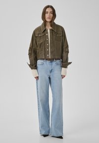 Femeie purtând o jachetă scurtă de piele verde măsliniu, mâneci de pulover crem, blugi cu talie înaltă, largi, de culoare albastru deschis și pantofi negri cu vârf ascuțit.