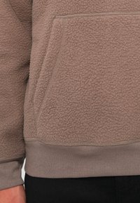 Close-up of a brown fleece sweater with a textured pocket and ribbed waistband worn over black pants.