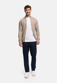 Beige kabelstrikket cardigan med glidelås foran, brukt over en hvit T-skjorte og marineblå bukser. Kombinert med hvite joggesko.