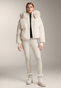Pujsasta jakna v kremni barvi s kapuco obloženo s krznom, sprednjimi gumbi in rebrastimi manšetami, kombinirana z ujemajočimi pajkicami in teksturiranimi belimi škornji.