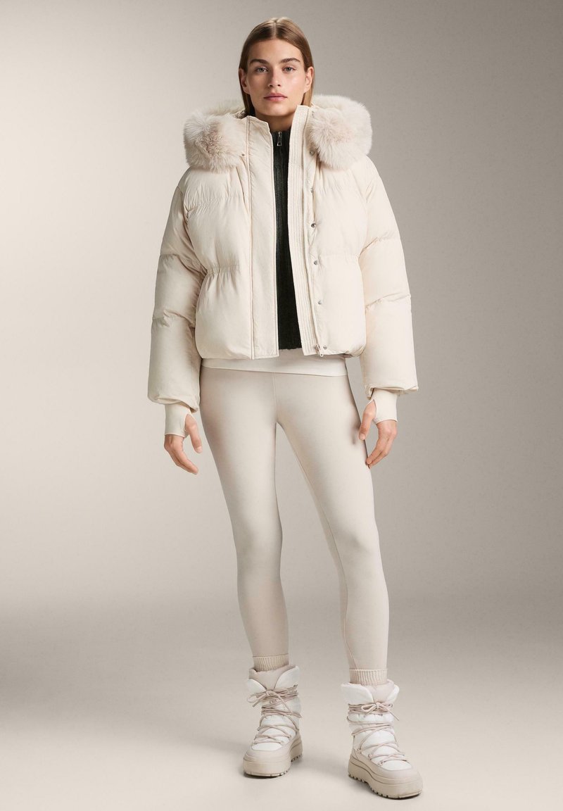 Pujsasta jakna v kremni barvi s kapuco obloženo s krznom, sprednjimi gumbi in rebrastimi manšetami, kombinirana z ujemajočimi pajkicami in teksturiranimi belimi škornji.