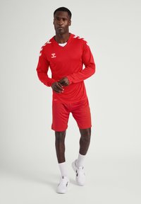 Rode sporttrui met lange mouwen en bijpassende shorts. Beschikt over witte accentstrepen op de schouders en een logo op de borst. Lichtgewicht stof.