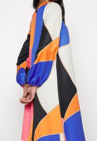 Femme en robe à manches longues avec des motifs géométriques abstraits en orange, bleu, noir, blanc et rose, debout les mains jointes dans le dos.