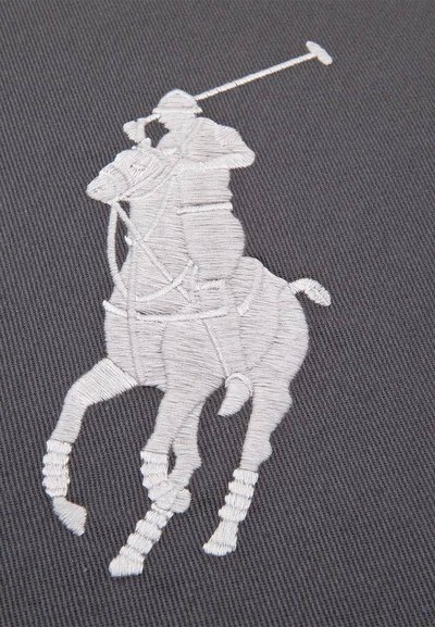 Λευκό κεντητό σχέδιο παίκτη πόλο που ιππεύει άλογο με σηκωμένο ρόπαλο σε σκουρόγριφο υφασμάτινο ύφασμα με υφή.