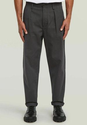 Hommes portant des pantalons plissés gris foncé et des bottes noires à lacets, debout avec les mains détendues le long du corps.