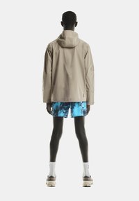 Beige waterdichte jas met capuchon, losse pasvorm. Gecombineerd met blauwe tie-dye shorts, witte sokken en lichtgekleurde schoenen. Achteraanzicht.