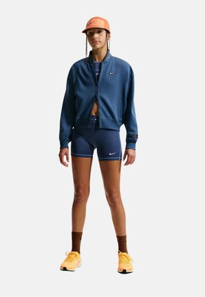 Mujer con chaqueta azul con cremallera, pantalones cortos de ciclismo azul marino a juego, gorra naranja, calcetines marrones y zapatillas de correr naranjas, de pie frente a un fondo blanco.