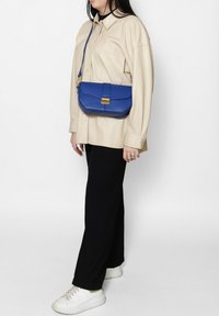 Sac à bandoulière en cuir bleu avec un fermoir doré, présentant un design structuré, une texture lisse et une longue sangle réglable.