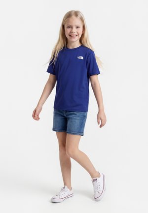 Junges Mädchen lächelt, trägt ein blaues Kurzarmshirt, Jeansshorts und weiße Sneakers, steht vor einem einfarbigen weißen Hintergrund.