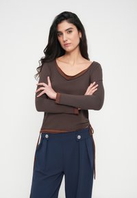 Καφέ μακρυμάνικο τοπ με στρογγυλή λαιμόκοψη, διακοσμημένο με ανοιχτόχρωμη καφέ λεπτομέρεια. Συνδυασμένο με ψηλόμεσο μπλε σκούρο παντελόνι με λεπτομέρειες κουμπιών.