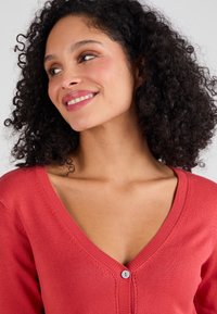 Koraalroze cardigan van zachte breiwerk, met een V-hals, iets getailleerd ontwerp en een lichtblauwe knop in het midden van de voorkant.