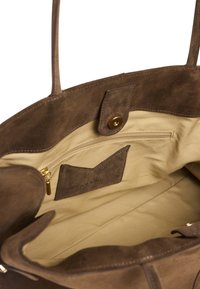 Sac cabas en daim marron avec un intérieur en tissu beige, doté d'une poche zippée et d'un patch logo. Comprend des bretelles d'épaule solides et une fermeture à pression magnétique.