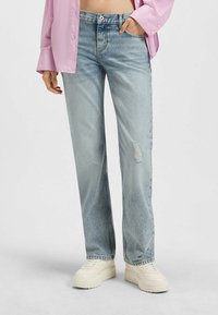 Ljusblå raka jeans med en slitna lapp på höger knä, bärs med en rosa skjorta och krämfärgade klumpiga sneakers.