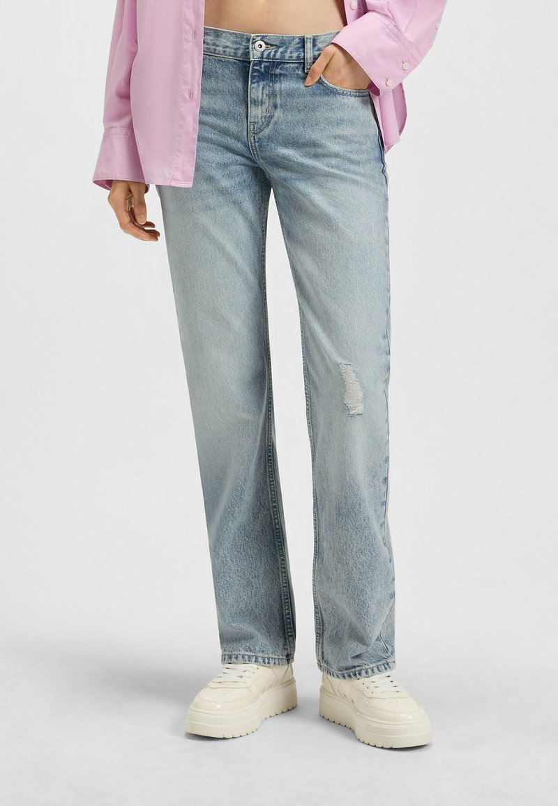 Ljusblå raka jeans med en slitna lapp på höger knä, bärs med en rosa skjorta och krämfärgade klumpiga sneakers.