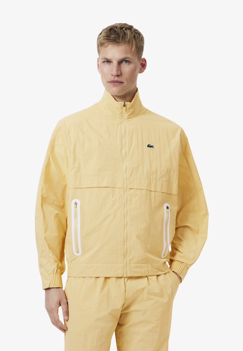 Man met kort blond haar die een gele jas met rits draagt en bijpassende broek met witte ritstaspockets en een klein logo op de borst.