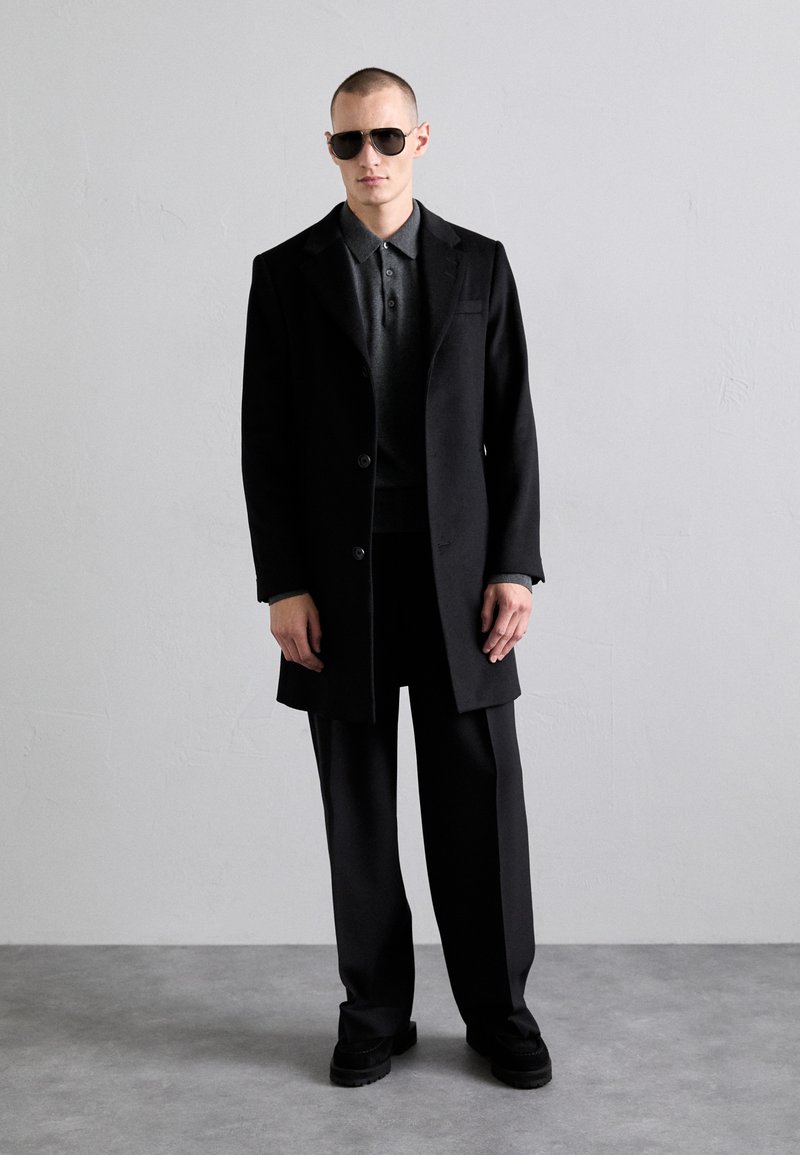 Manteau en laine noir sur un polo sombre, avec un pantalon ample et des chaussures noires. Le modèle porte des lunettes de soleil, et l'arrière-plan est neutre.