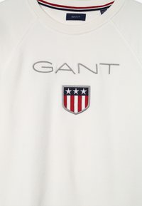 Witte sweatshirt met een grijs geborduurd "GANT" logo en een schild met rode en blauwe strepen en drie sterren, voorzien van een geribde kraag.