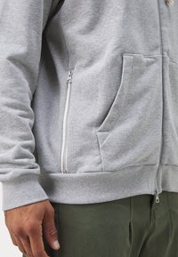 Homme portant un sweat à capuche gris clair zippé avec poches avant et pantalon vert, main détendue visible sur le côté.