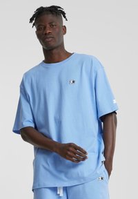 Light blue cotton t-shirt with short sleeves, round neckline, and a small black logo on the left side. Plain design, relaxed fit.