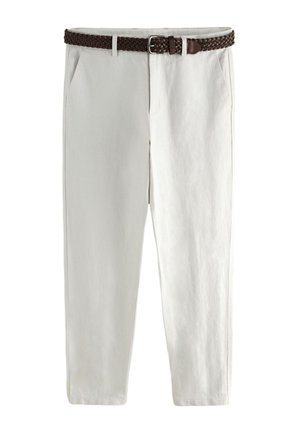 Homme debout, mains dans les poches, portant une chemise beige à manches longues, un pantalon blanc, une ceinture noire et des sandales noires sur fond gris.