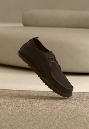 Dunkelbrauner, lässiger Schnürschuh schwebt über einem strukturierten beigen Teppich mit einem beigefarbenen Sofa im Hintergrund.