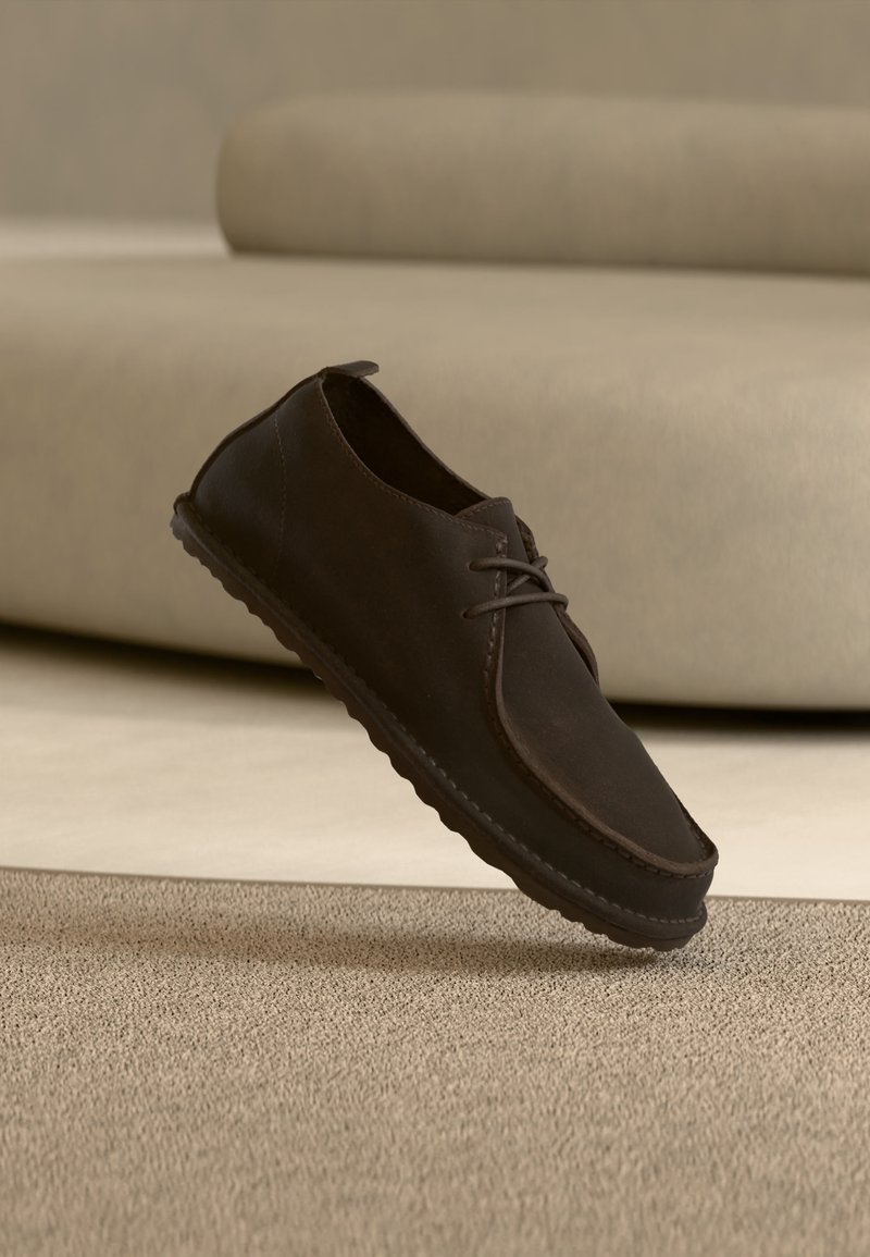 Σκούρο καφέ καθημερινό παπούτσι με κορδόνια που αιωρείται πάνω από μια υφασμάτινη μπεζ μοκέτα με έναν μπεζ καναπέ στο φόντο.