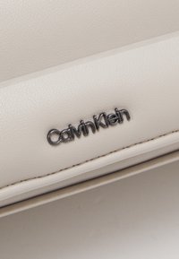 Logo Calvin Klein na teksturowanym, kremowym materiale o gładkim wykończeniu. Logo jest w eleganckim czarnym kroju pisma, wyśrodkowane na produkcie.