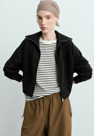 Jeune femme portant un foulard à carreaux, une veste noire zippée, une chemise rayée blanche et noire, et un pantalon marron, les mains sur les hanches.