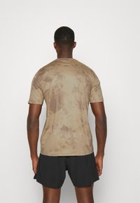 Hnědé tričko s krátkým rukávem a texturovaným, žíhaným vzorem. Má kulatý výstřih a tři malé tečky na zadní straně u límce.