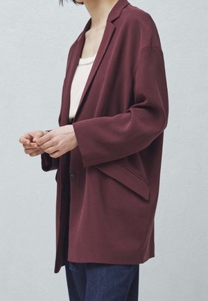 Osoba ubrana w luźno dopasowaną bordową marynarkę na białym topie i ciemnoniebieskich spodniach, dłonie delikatnie złożone z przodu.