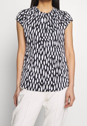 Femme portant un chemisier sans manches à motif noir et blanc avec de petits plis au niveau de l'encolure et un pantalon blanc.