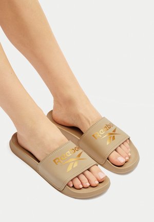 Beżowe klapki Reebok z szerokim paskiem z złotym logo, teksturowaną wkładką i płaską podeszwą. Idealne dla komfortu i łatwego noszenia.