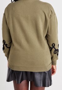 Oliwkowa zielona bluza z czarnymi haftowanymi akcentami na rękawach, ściągaczami przy mankietach i u dołu oraz luźnym krojem. Nosi się ją na czarną skórzaną spódnicę.