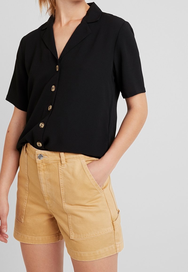Donna che indossa una camicia nera a maniche corte con bottoni, infilata in pantaloni shorts beige a vita alta con tasche frontali, mano in tasca, su uno sfondo neutro.