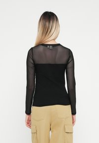 Musta pitkähiha shirt, jossa on läpinäkyvä verkkotakaosa, kiinteä musta etuosa ja pyöreä kaula-aukko. Kangas on sileä ja siinä on hieman joustavuutta.