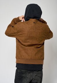 Veste en tissu texturé marron avec une capuche noire, des poignets côtelés et des coutures visibles. Dispose d'un panneau arrière avec des perforations et d'une poche inférieure.
