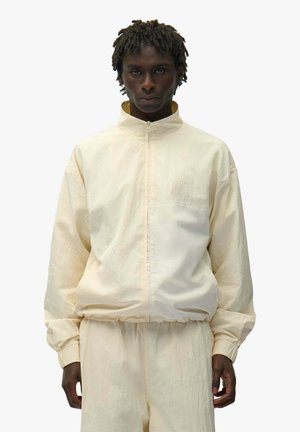 Uomo dalla pelle scura e capelli ricci corti che indossa una giacca crema con cerniera ampia e pantaloni abbinati, in piedi davanti a uno sfondo semplice.