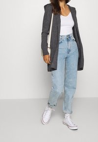 Grå stickad cardigan med huva, över en vit V-ringad topp, kombinerad med ljusblå högmidjade jeans och vita högstövelsneakers.