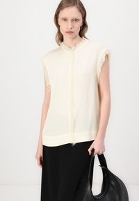 Light yellow sleeveless top with fringe detailing along the collar and armholes, paired with a black bag. Smooth fabric texture.