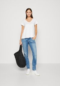 Weißes V-Ausschnitt-T-Shirt, hellblaue Jeans und weiße Sneakers. Hält eine schwarze geflochtene Tragetasche. Einfaches, lässiges Outfit vor einem neutralen Hintergrund präsentiert.