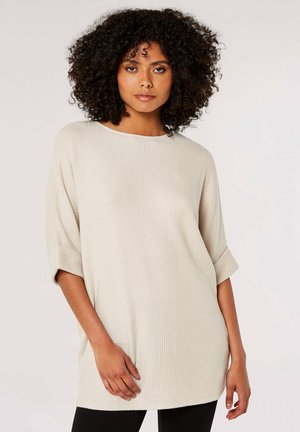 Light beige oversized knit top with a ribbed texture, round neckline, and elbow-length sleeves. Worn with fitted black leggings.